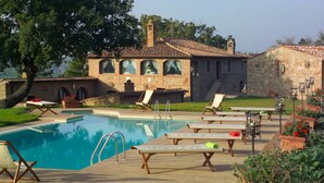Ein Poolbereich mit Liegestühlen, ein Gebäude aus Stein mit Rundbogenfenstern und ein klares, blaues Schwimmbecken.