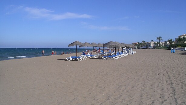 A beach with rows of sun loungers and thatched umbrellas, a clear sky, and the sea in the background. - 117 Las Velas, 57 (Denia)