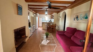 A spacious living area with a red sofa, a glass coffee table, and a fireplace. A kitchen is visible in the background with a dining table and chairs. - 117 Las Velas, 57 (Denia)