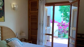 A bedroom with a bed, a ceiling fan, a nightstand, a lamp, and a view of the outside balcony with flowers. - 117 Las Velas, 57 (Denia)