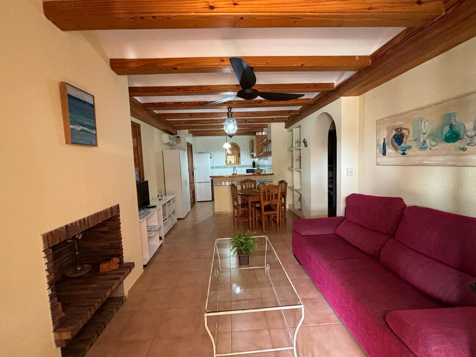 A living room with a red sofa, a glass coffee table, and a fireplace. There is a kitchen in the background with wooden beams on the ceiling.
