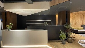A modern reception area with a wooden reception desk, potted plants, and a dark wall with horizontal light strips. - R Hotel Namba (Osaka)