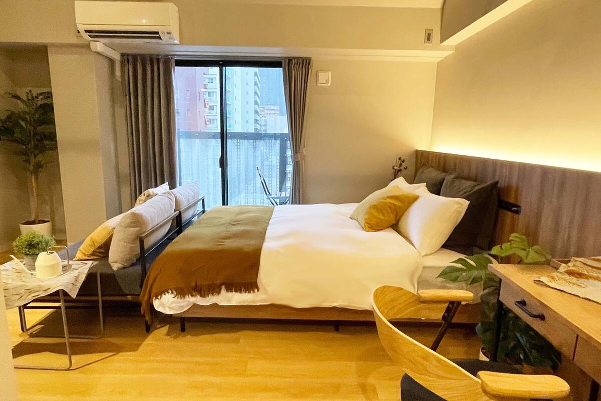 A hotel room with a bed, a desk, a chair, a potted plant, and a window with curtains.
