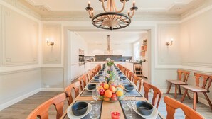 A long dining table set for a meal with chairs on both sides, a chandelier above, and a kitchen in the background. - Ecolodge in Remouchamps with sauna (Remouchamps)