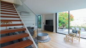 Um interior moderno com escada de madeira, TV de tela plana, mesa de centro redonda e vista para um jardim através de grandes janelas.