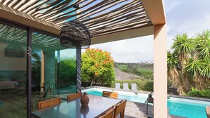 Uma área de refeições moderna ao ar livre, com mesa e cadeiras de vidro, um vaso decorativo e vista para uma piscina e paisagem.