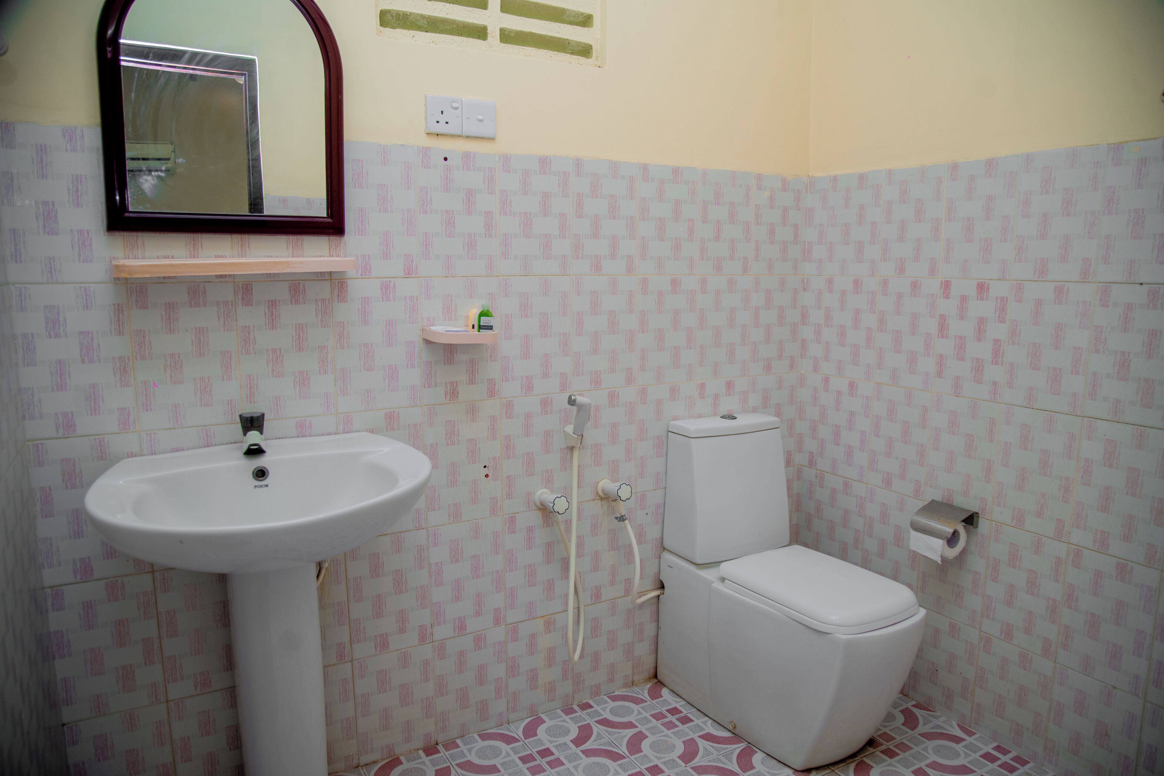 Un baño con lavamanos, inodoro y ducha blancos. Hay un espejo sobre el lavamanos y una repisa con artículos de aseo.