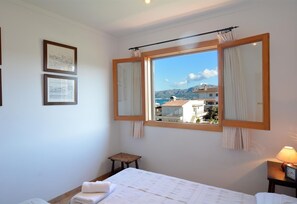 A bedroom with a large window, a bed, a small stool, and a nightstand with a lamp. - Apartment 'Calabruix' with Sea View, Wi-Fi and Air Conditioning (Son Serra de Marina)