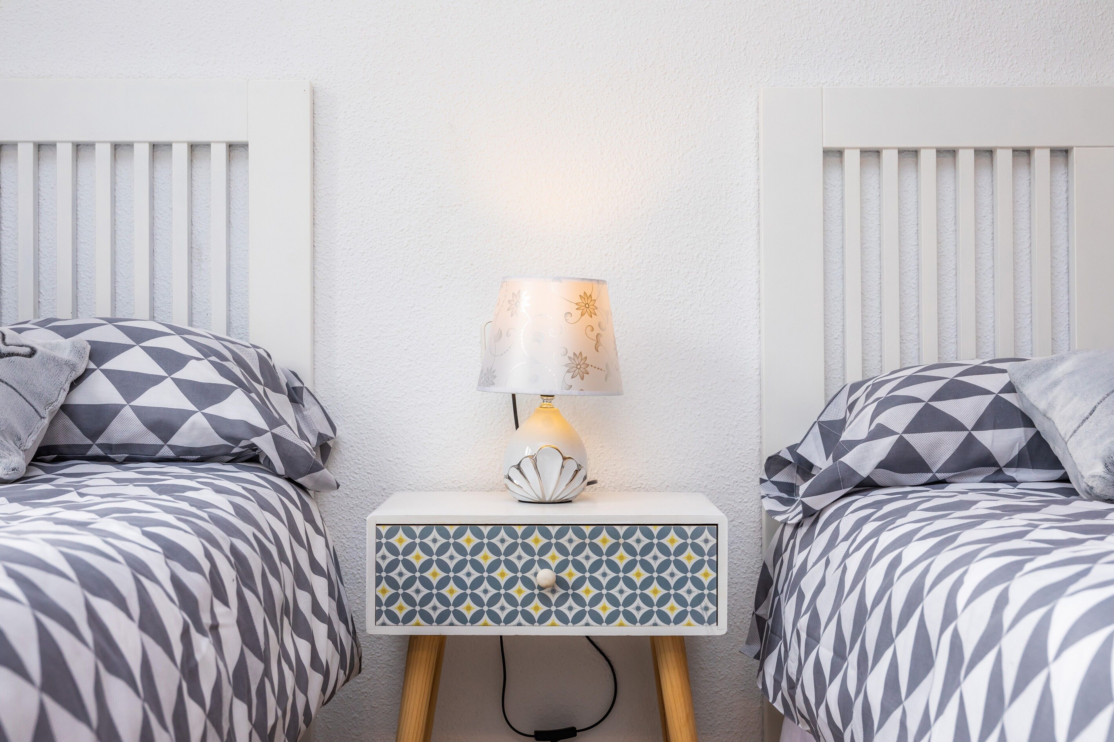 A bedroom with two beds, a bedside table with a lamp, and a patterned drawer unit.