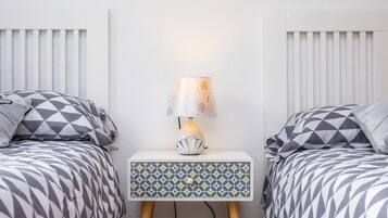 A bedroom with two beds, a bedside table with a lamp, and a patterned drawer unit.