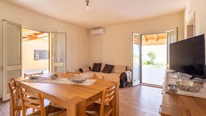 A dining area with a wooden table and chairs, a sofa, and a television in a room with an open layout and wooden flooring. - Holiday Home "Casa Vacanza Il Melograno" with 2 Private Terraces, A/C and Wi-Fi (Santa Maria del Focallo)