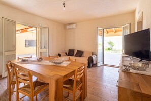A dining area with a wooden table and chairs, a sofa, and a television in a room with an open layout and wooden flooring.