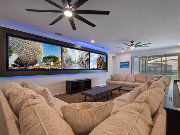 A spacious living room with a large sectional sofa, a flat-screen TV mounted on the wall, and a ceiling fan.