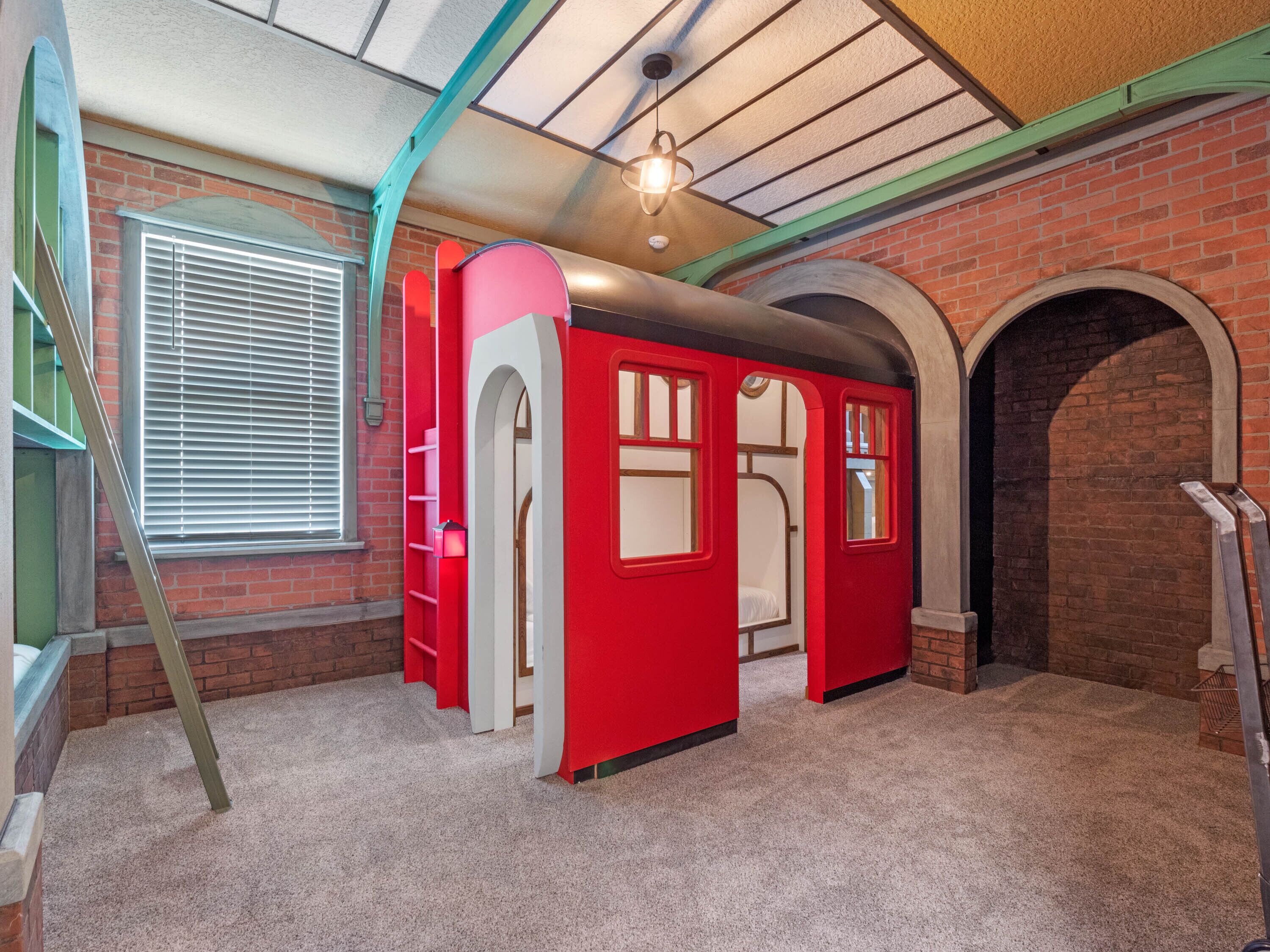 A room with a brick wall, a window with blinds, a red playhouse, and a ladder.
