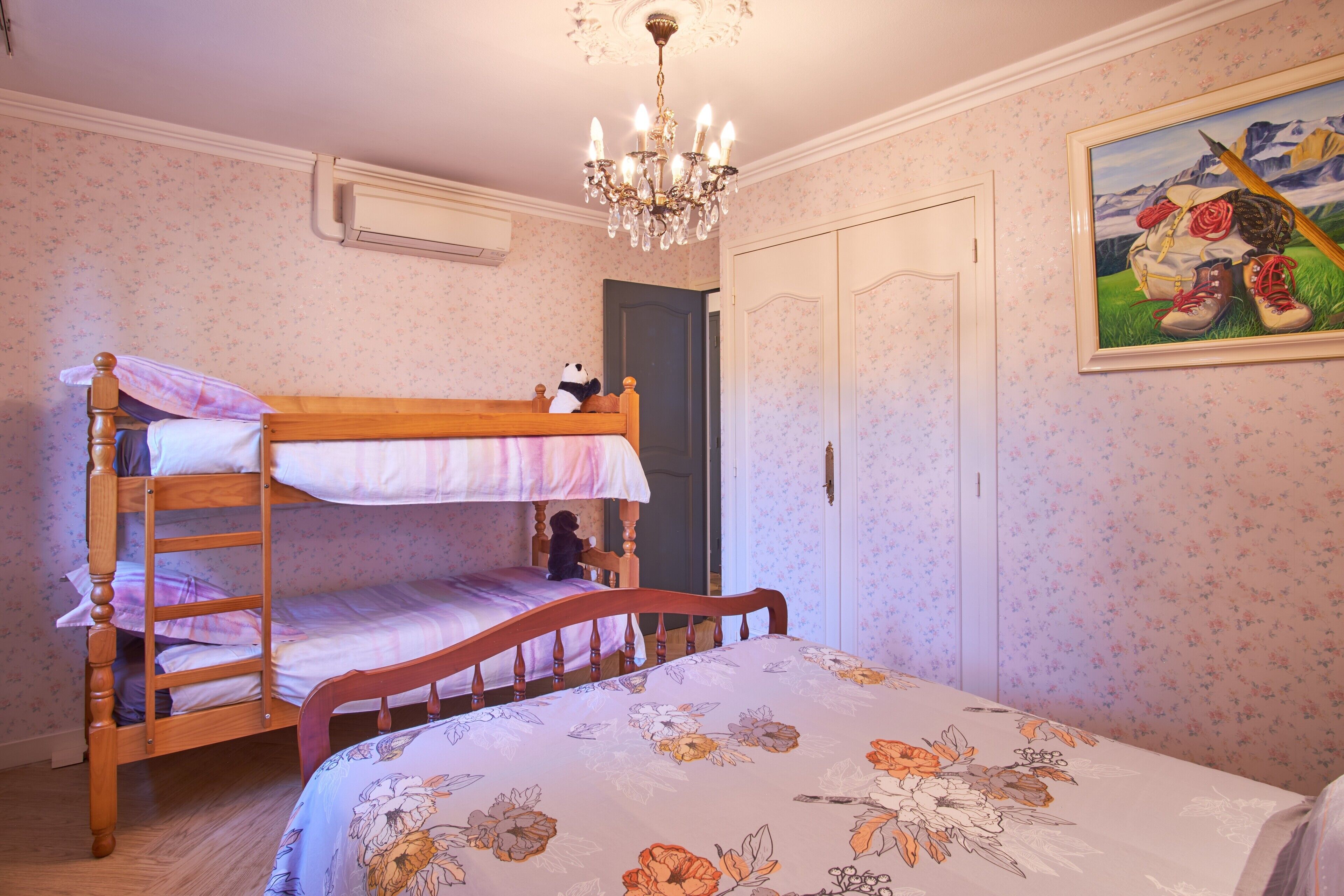 Una camera da letto con un letto a castello, una sedia di legno, un lampadario, una porta e una coperta con motivo floreale.