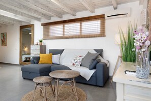 A modern living room with a grey sofa, two round wicker stools, a wooden ceiling, and a large window with blinds.