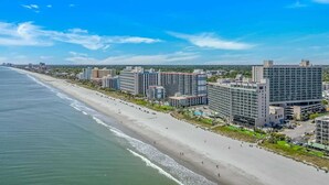 Een kuststadsgezicht met een hotel aan het strand, met meerdere hoge gebouwen, een zandstrand en een helderblauwe lucht.