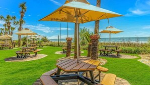 Een terras aan het strand met houten tafels en parasols, palmbomen en uitzicht op de oceaan.