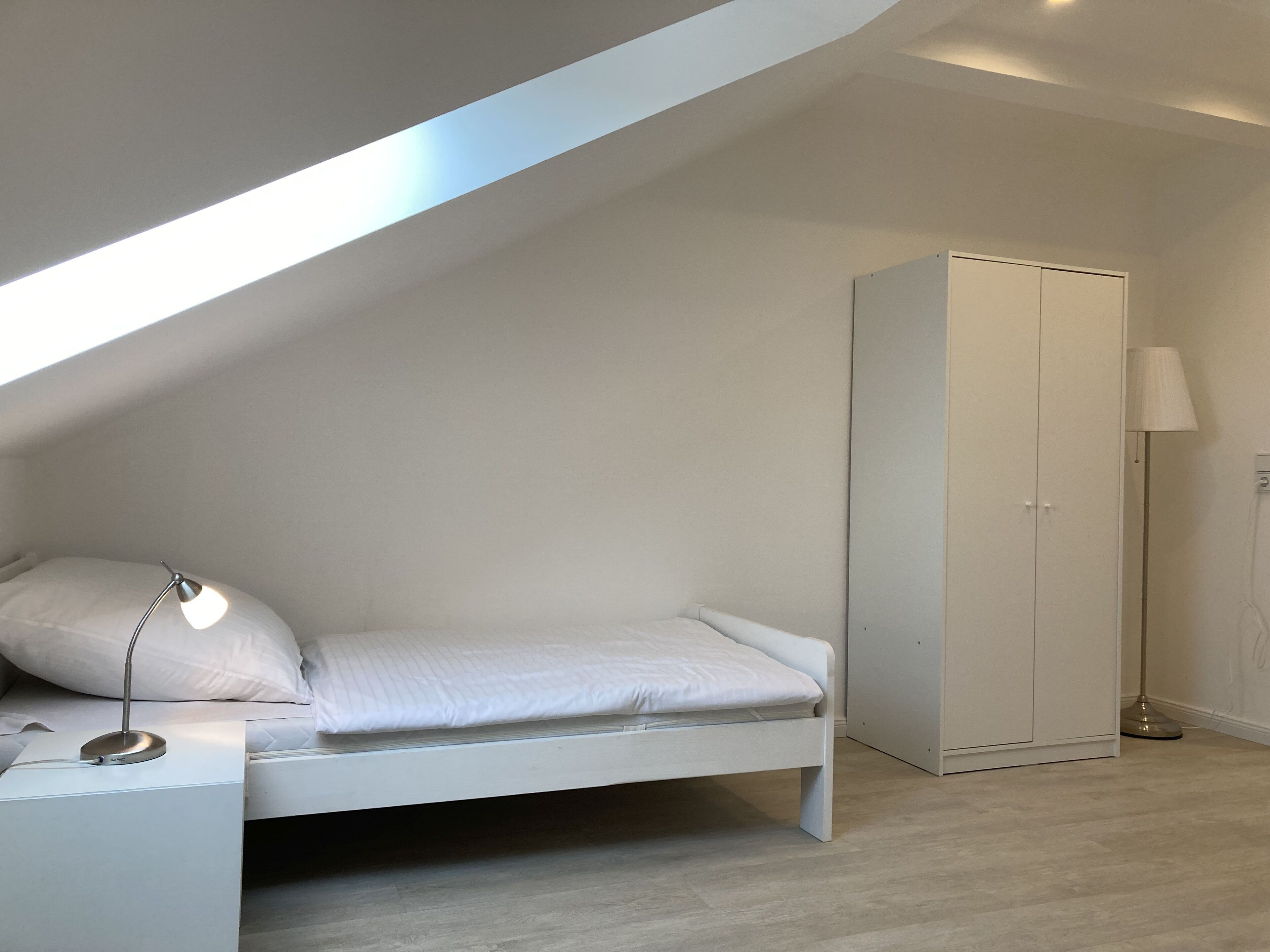 Une chambre avec un plafond en pente, un lit simple, une armoire et une lampe sur pied.