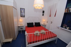 A bedroom with a red bedspread, white headboard, bedside tables with lamps, a wardrobe, and a small step stool. - Dandy Villas Velanidia Sailor's House (Monemvasia)