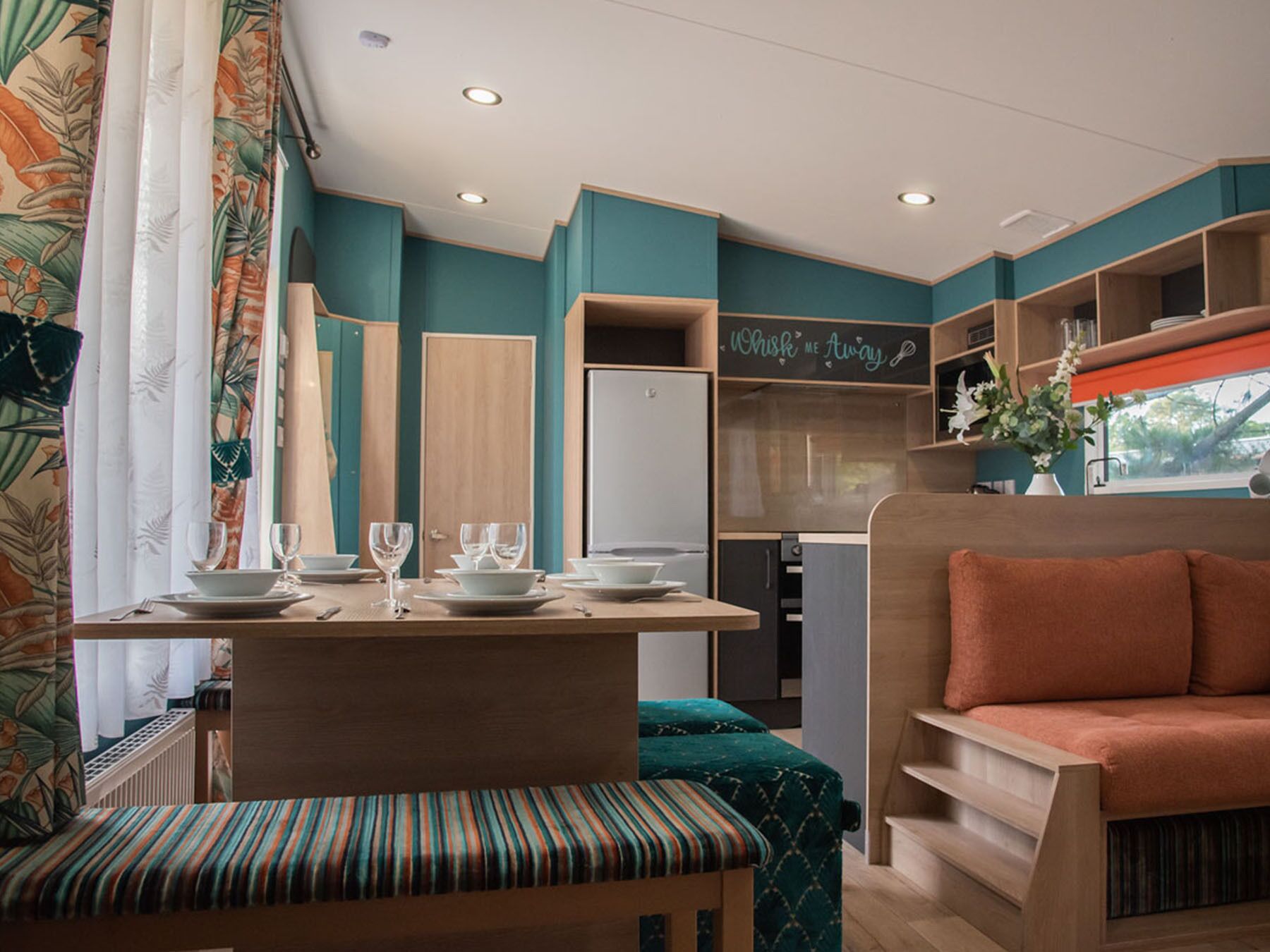Eine kompakte, moderne Küchenzeile mit türkisfarbenen Wänden, einem Holztisch gedeckt mit Tellern und Gläsern, und einer gemütlichen Sitzecke mit gepolstertem Sitzbrett und Sofa.