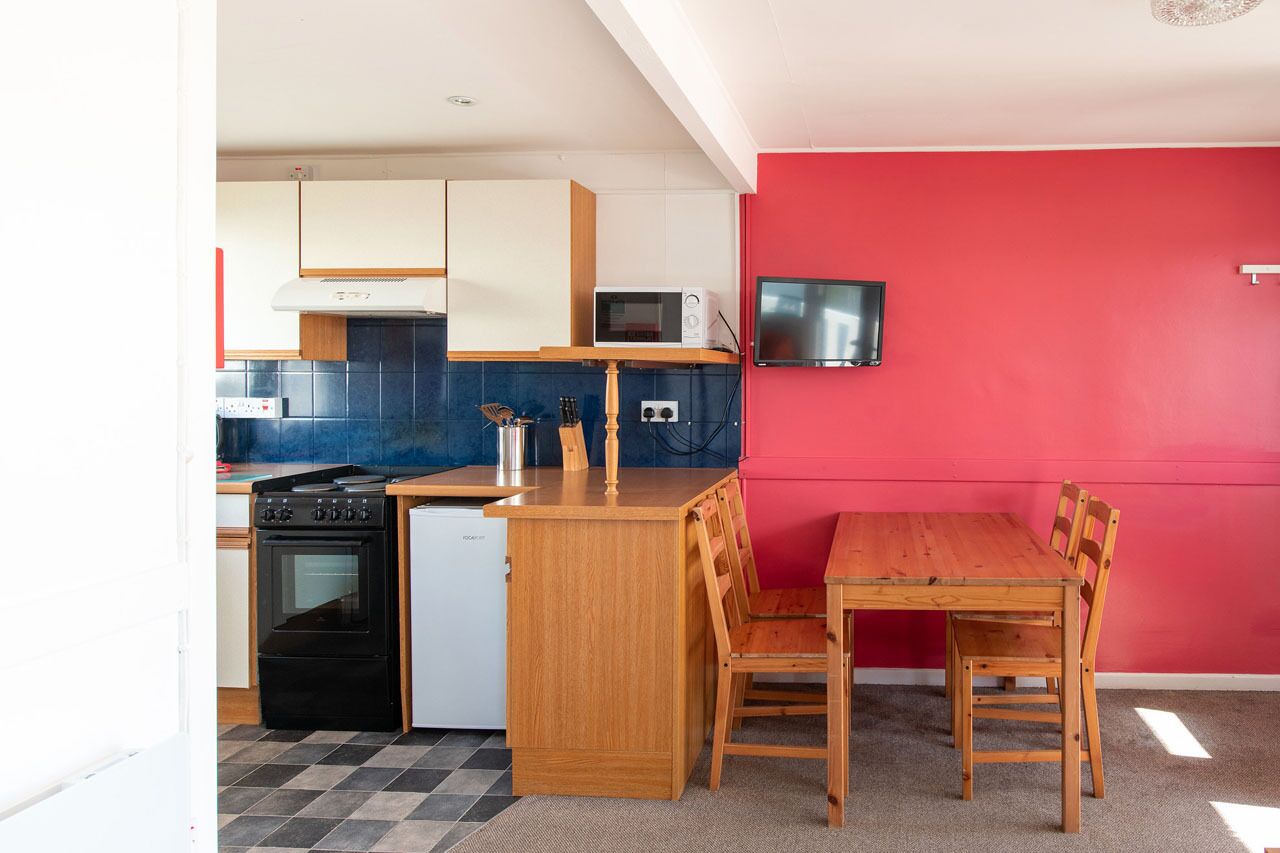 Een praktische keuken met een magnetron, fornuis en vaatwasser, een eettafeltje met stoelen, en een tv bevestigd aan een rode muur.