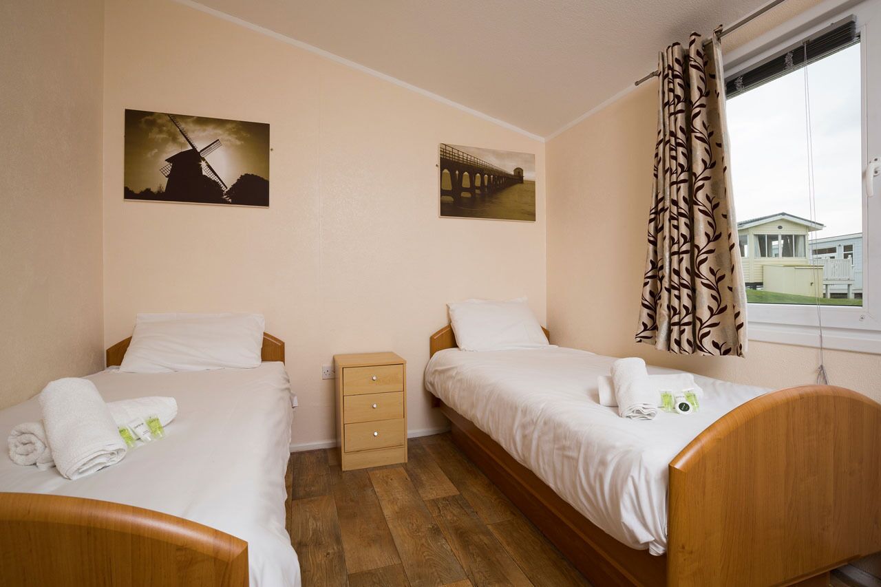 A hotel room with two single beds, a bedside table with a lamp, a window with curtains, and two wall-mounted pictures.