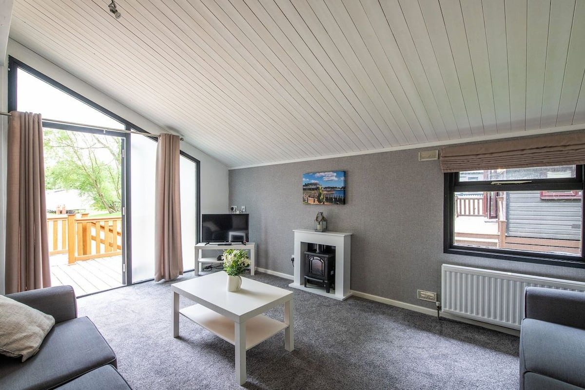 A modern living room with a fireplace, a television, and a large window overlooking a deck.