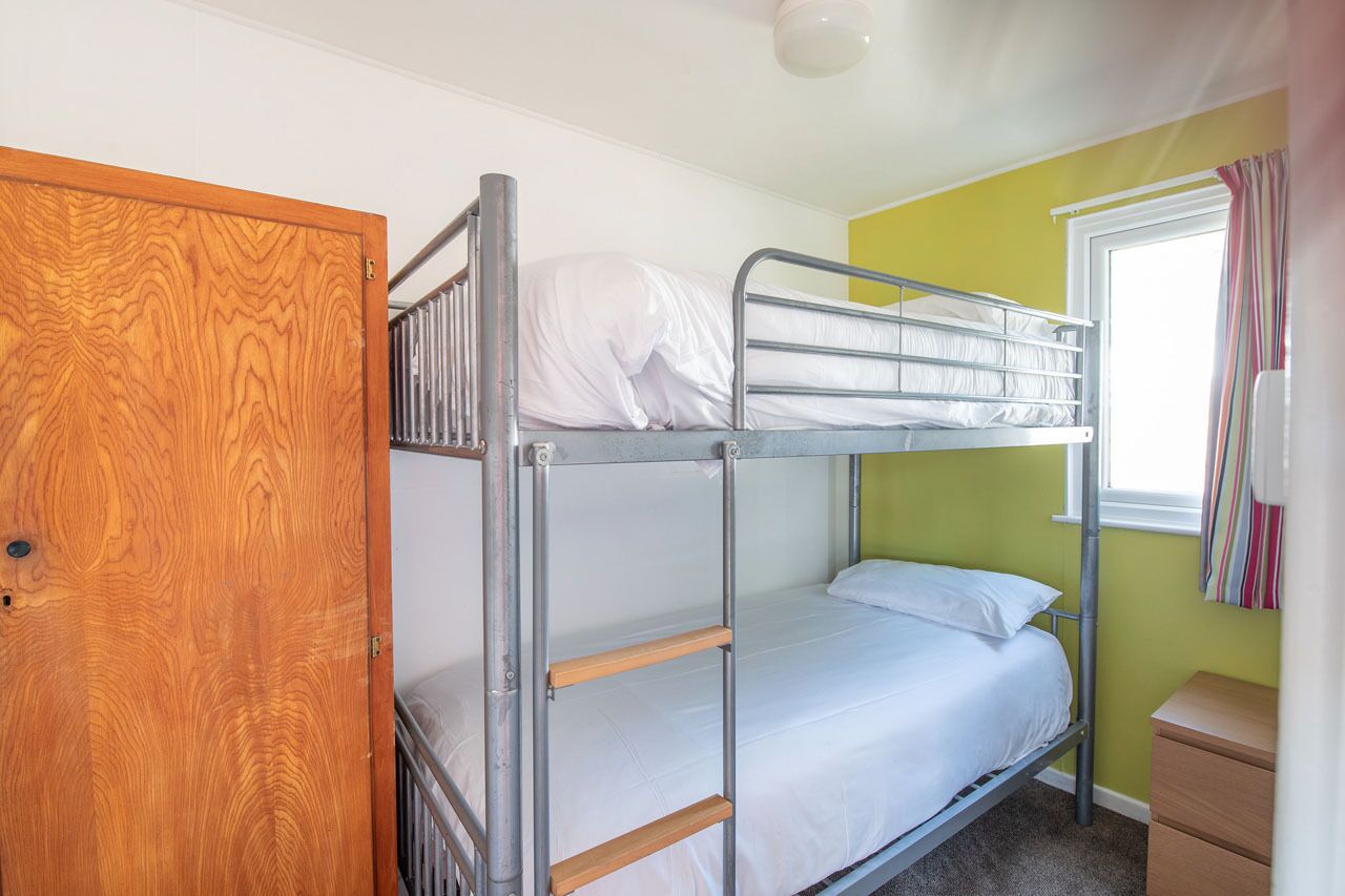 A bunk bed room with a wooden door, a window with curtains, and a small bedside table.