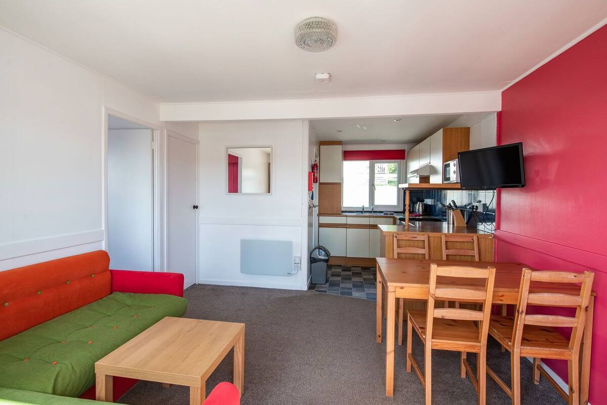 A compact living space with a green and red sofa, a wooden dining table, and a kitchen area with a TV.