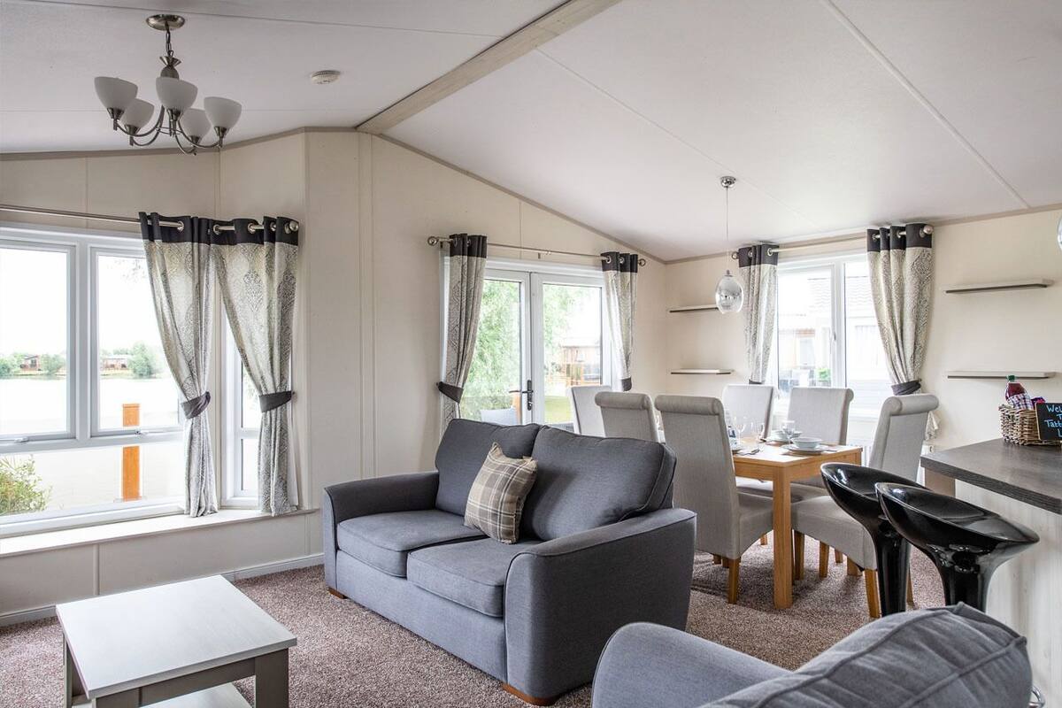 A modern living room with a grey sofa, dining table, and chairs. There are large windows with curtains, a chandelier, and a small table.