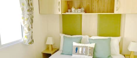 A bedroom with a bed, two bedside tables, a nightstand, a wooden headboard, and a shelf with decorative items.