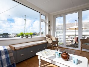 Een knus vertrek met een groot raam, een bankje, een tafel met een kaart en mokken, en een rieten stoel.