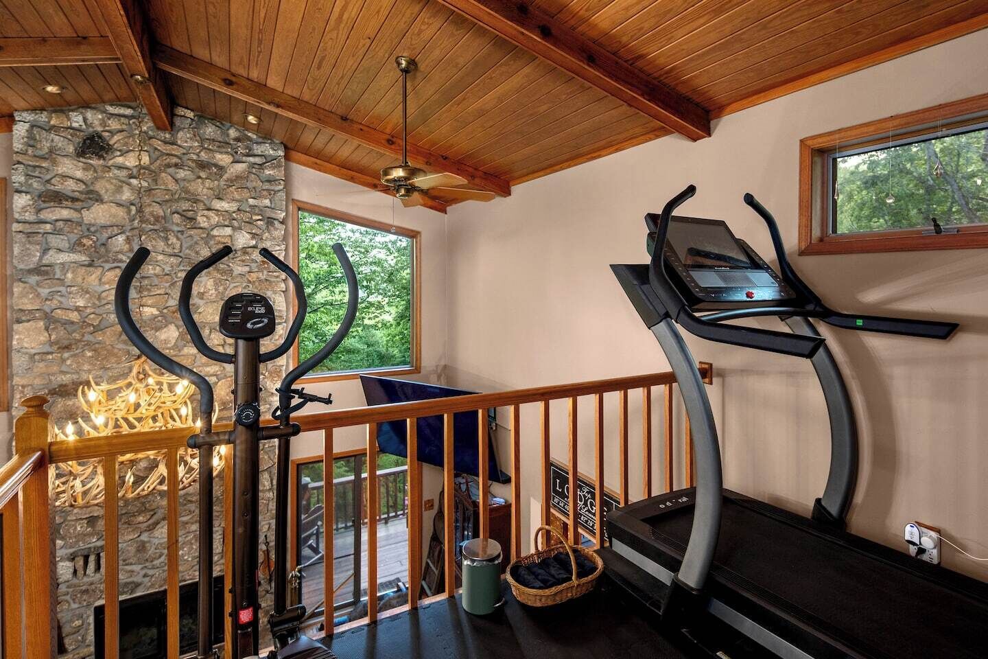 Une salle de sport à domicile équipée d’un tapis de course, d’une cheminée en pierre et d’une balustrade en bois.