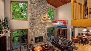 A spacious living room with a stone fireplace, large windows, a chandelier, and a television. - One of a kind, Creek Front Lodge 3 floors, 2 kitchens, long distance views. (MARS HILL)