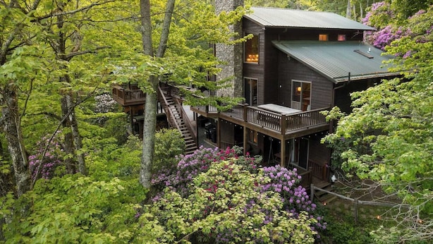 A cabin with a wooden deck and a stone chimney, surrounded by lush greenery and flowering bushes. - One of a kind, Creek Front Lodge 3 floors, 2 kitchens, long distance views. (MARS HILL)