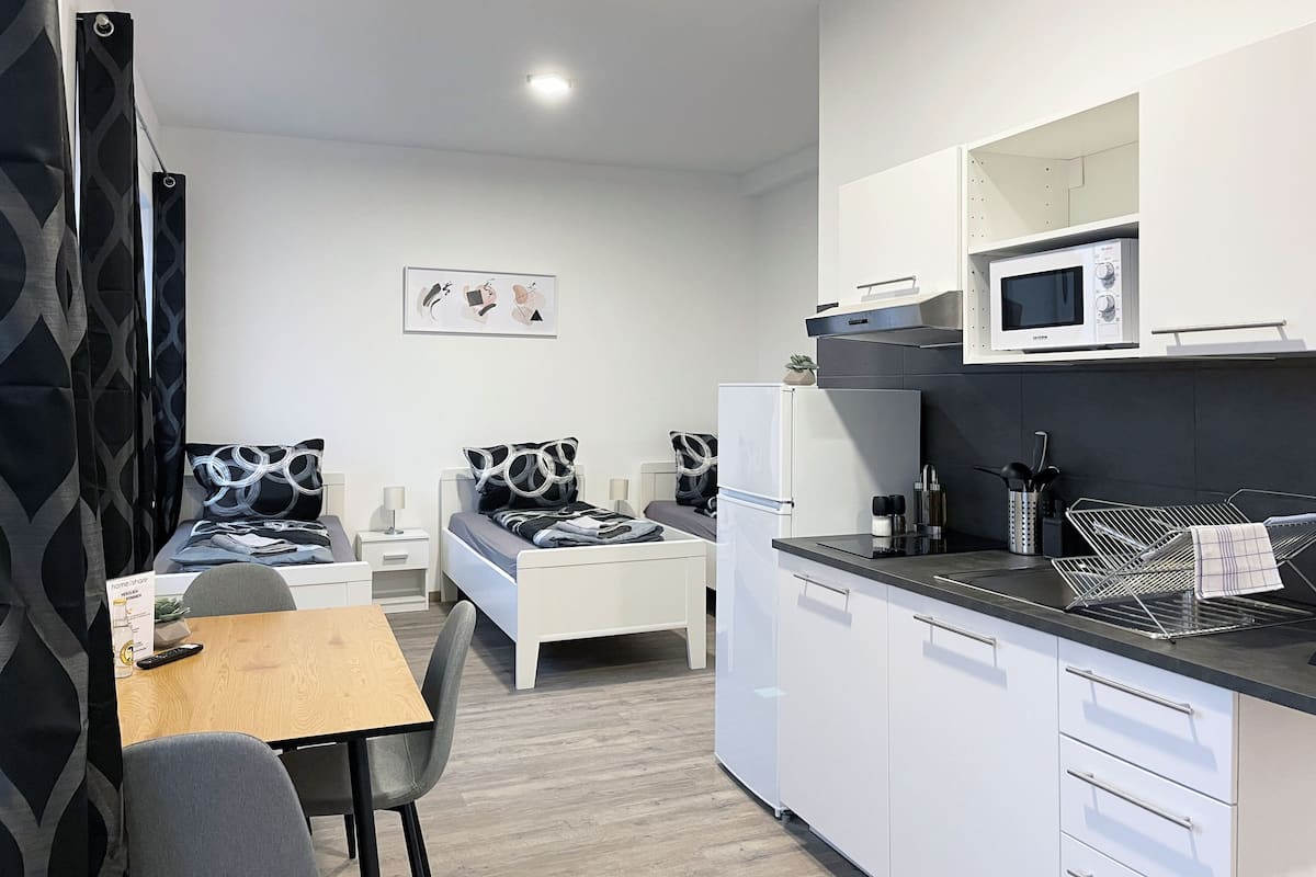 Een compacte hotelkamer met kitchenette, een bed met geborduurde kussens, een eettafeltje en een stoel.