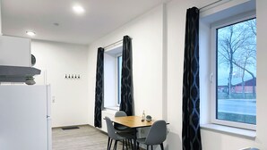 A modern room with a dining area, a bed, and a large window with curtains. - Hochwertige Studios für Monteure (Stemshorn)