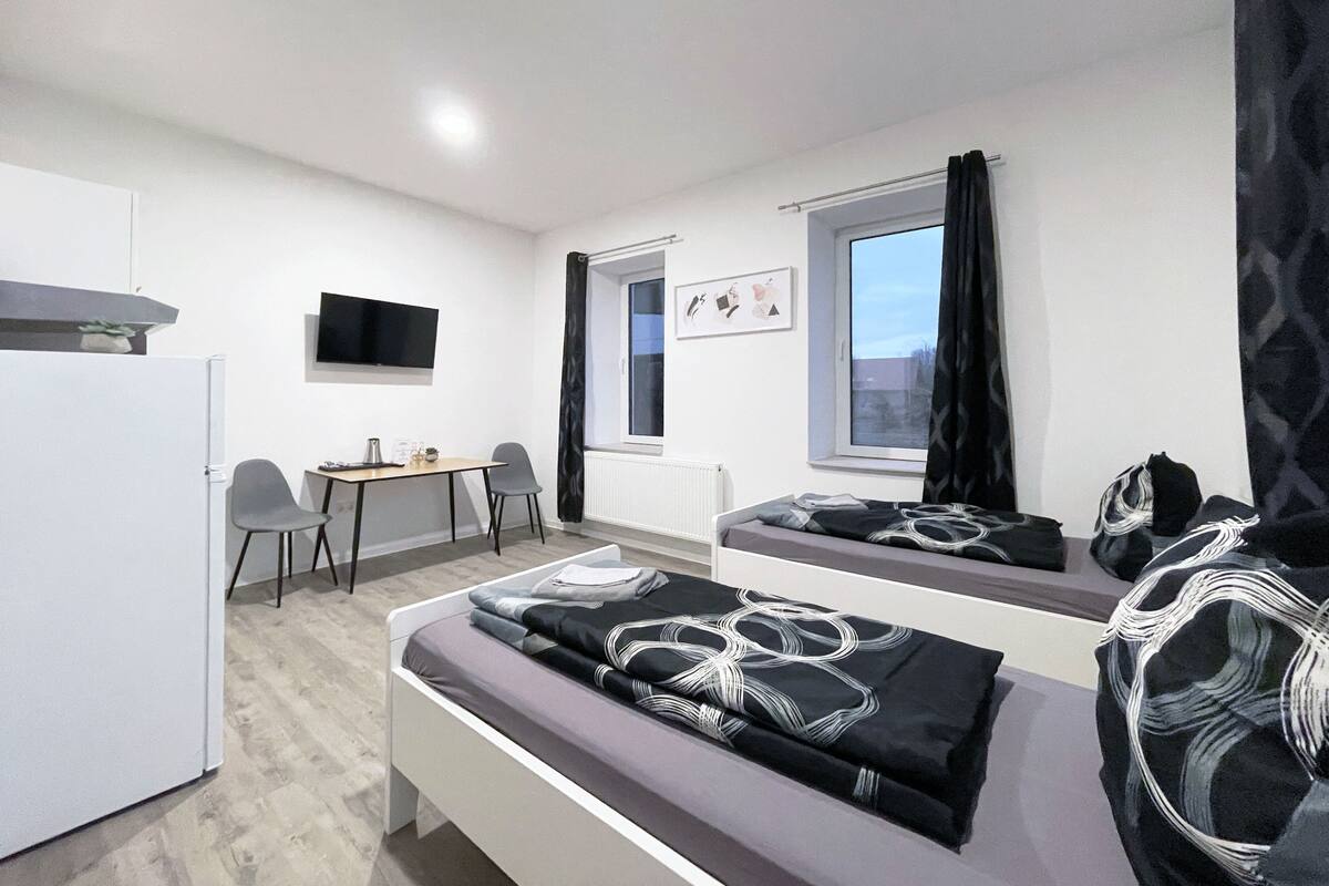 Een moderne slaapkamer met een bed, een bureau en een stoel. Er is een raam met gordijnen, een flatscreen televisie aan de muur en een radiator.