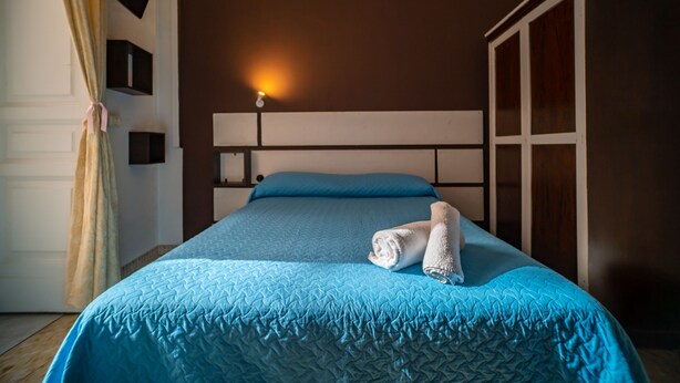 A neatly made bed with a blue quilt and pillows, a bedside lamp, and a wardrobe in a hotel room.