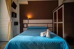 Ein ordentlich bezogenes Bett mit blauer Steppdecke und Kissen, eine Nachttischlampe und ein Kleiderschrank in einem Hotelzimmer.