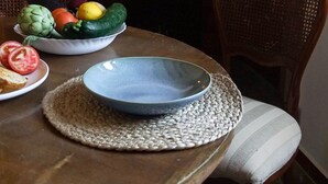 A dining table with a bowl of vegetables, a plate of sliced tomatoes, and a blue bowl. There are two wicker chairs around the table, and a patterned wall above the fireplace.