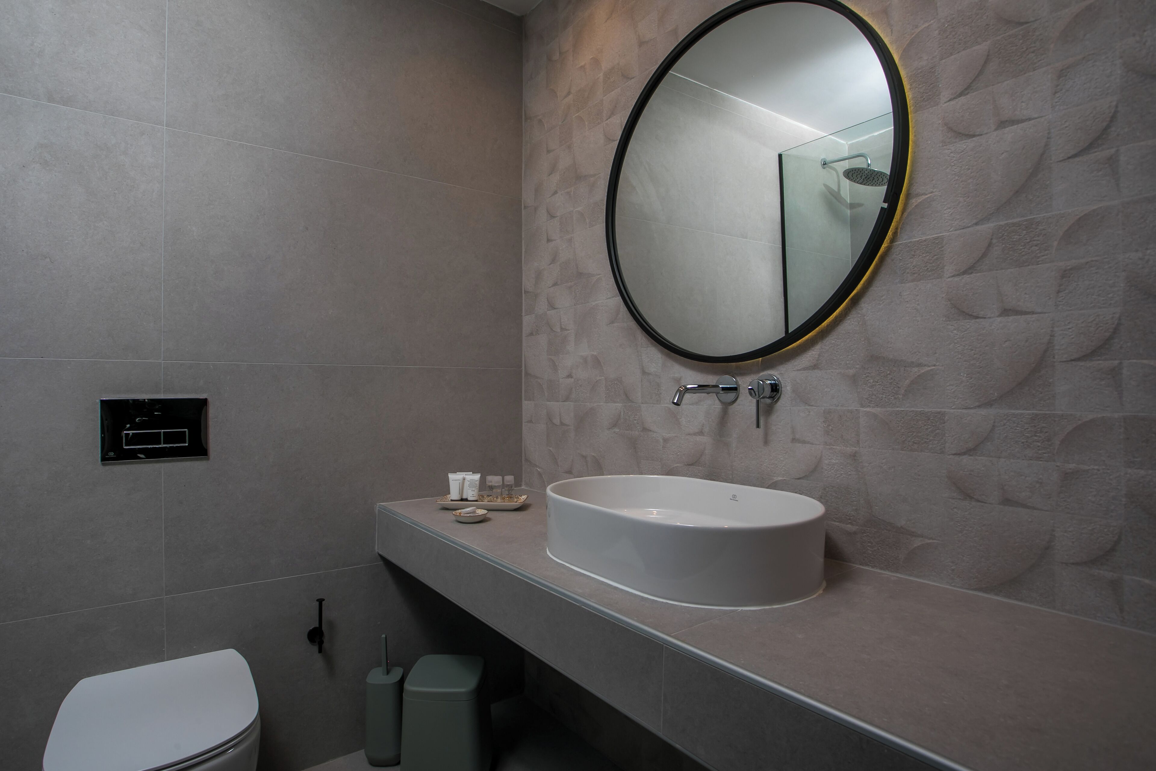 A modern bathroom with a large oval mirror, a white sink, and a toilet.