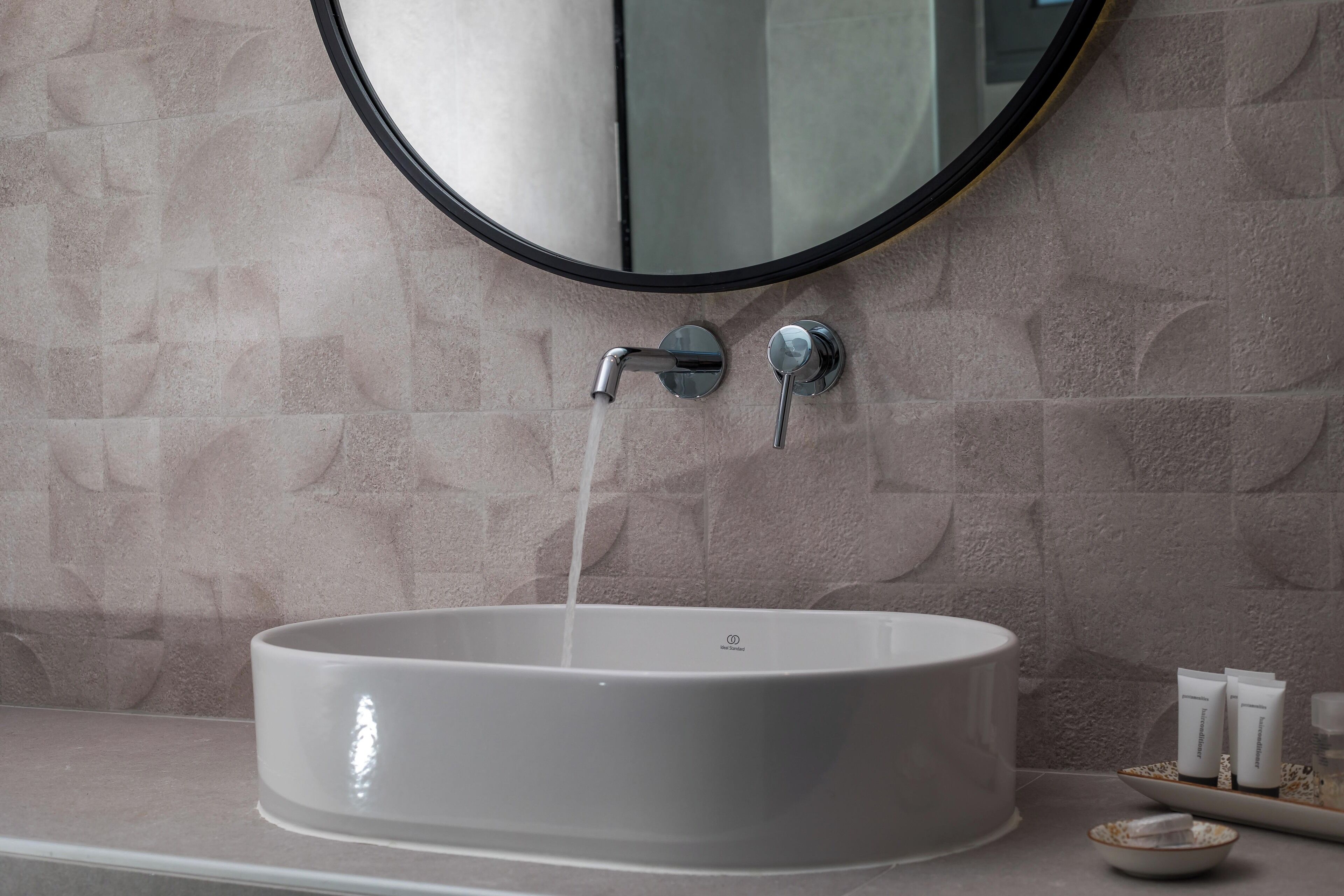 A bathroom with a round mirror, a sink with running water, and toiletries on the counter.