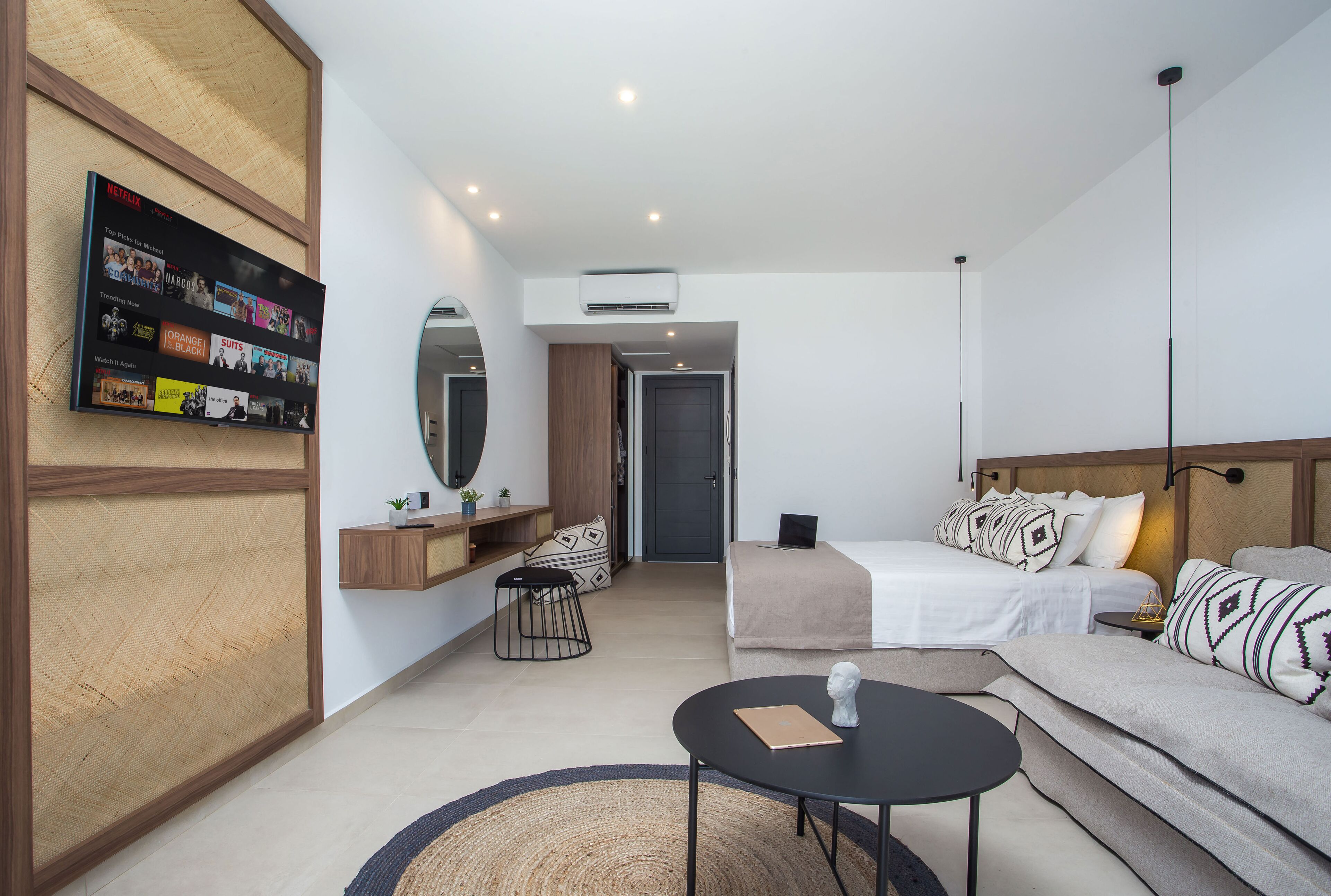 Una habitación de hotel moderna con una televisión de pantalla plana en la pared, una cama con edredón blanco, un sofá con cojines estampados, una mesita redonda y un espejo sobre el sofá.