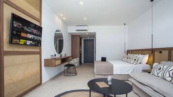 Una habitación de hotel moderna con una televisión de pantalla plana en la pared, una cama con edredón blanco, un sofá con cojines estampados, una mesita redonda y un espejo sobre el sofá.
