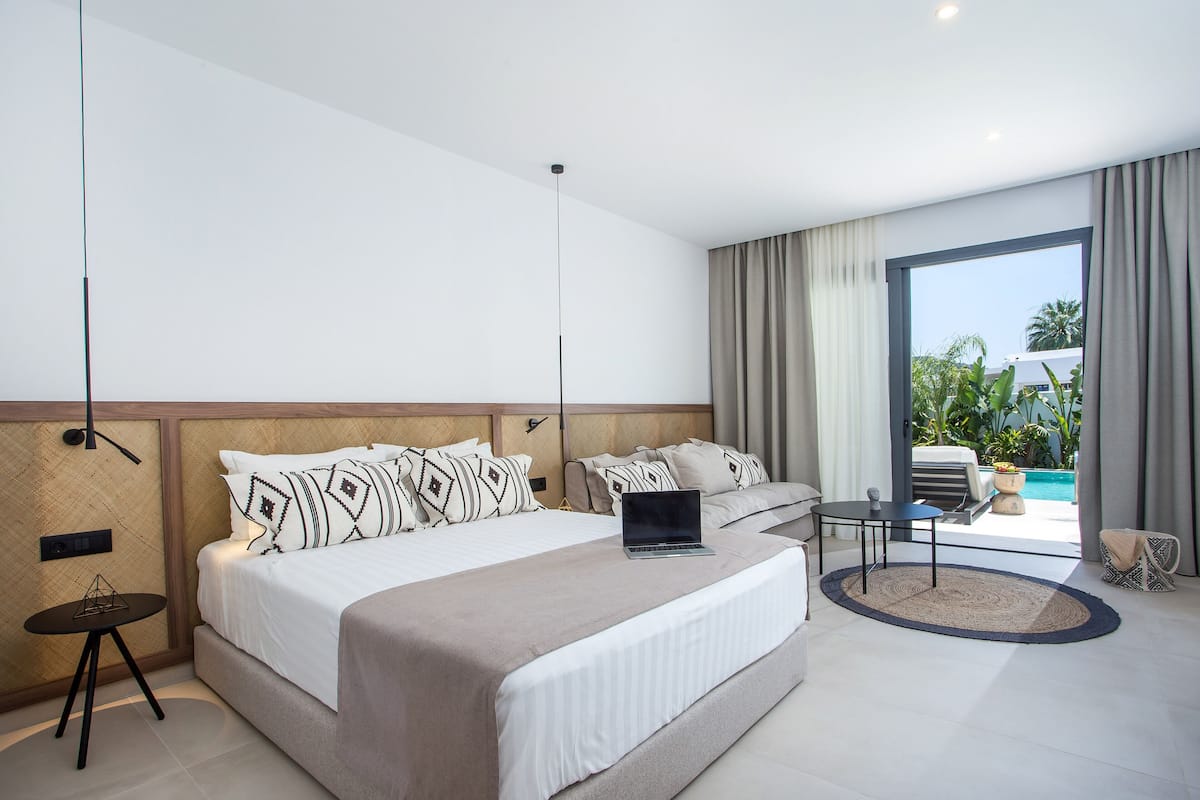 Una habitación de hotel moderna con una cama grande, un ordenador portátil sobre la cama, una mesa redonda y vistas a una piscina.