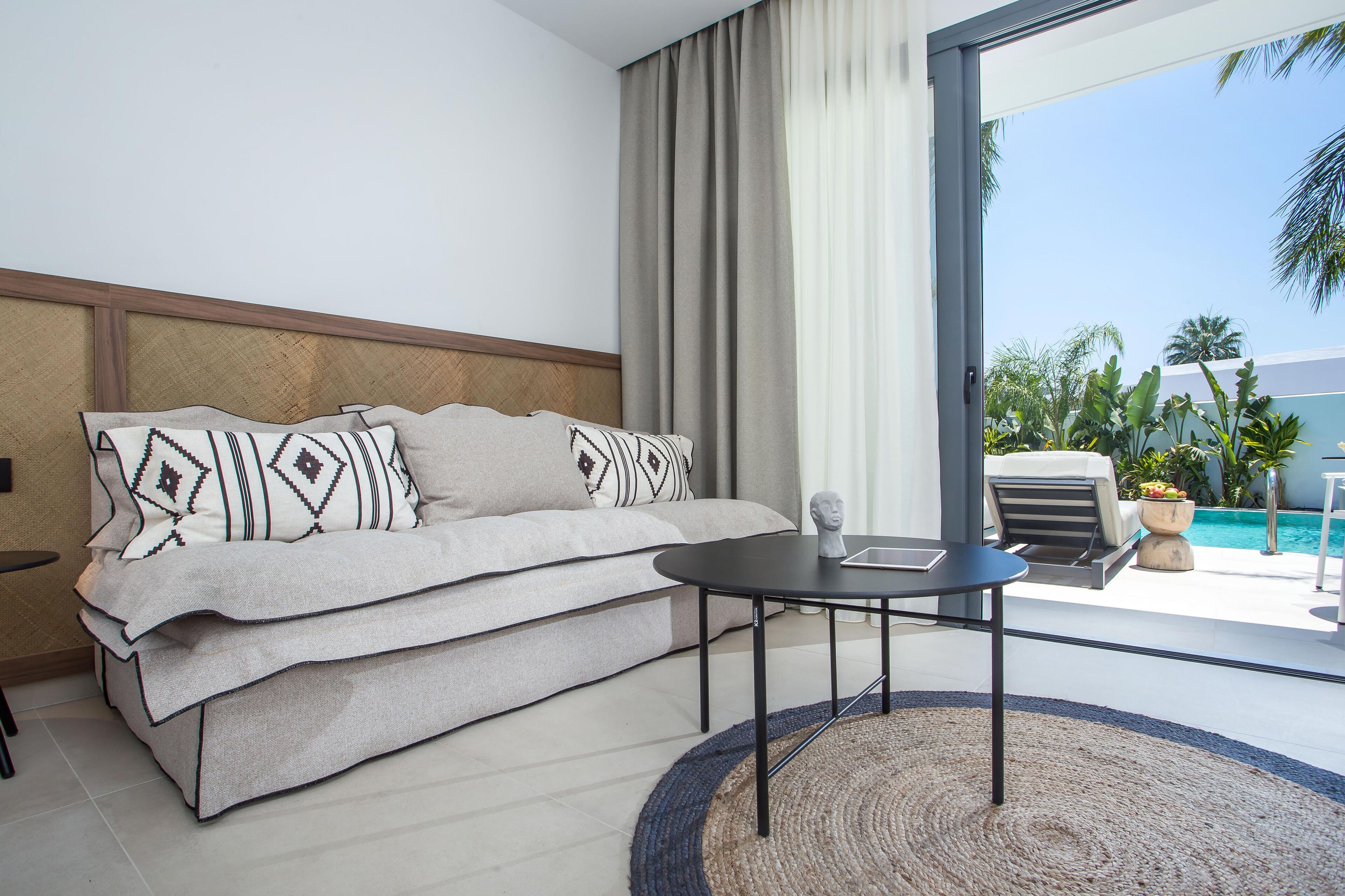 Habitación de hotel moderna con sofá, una mesita y vistas a una piscina y palmeras.