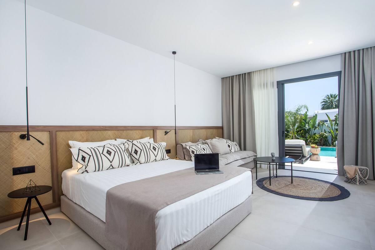 Habitación de hotel moderna con una cama grande, una laptop sobre la cama, una mesita redonda y vistas al exterior.