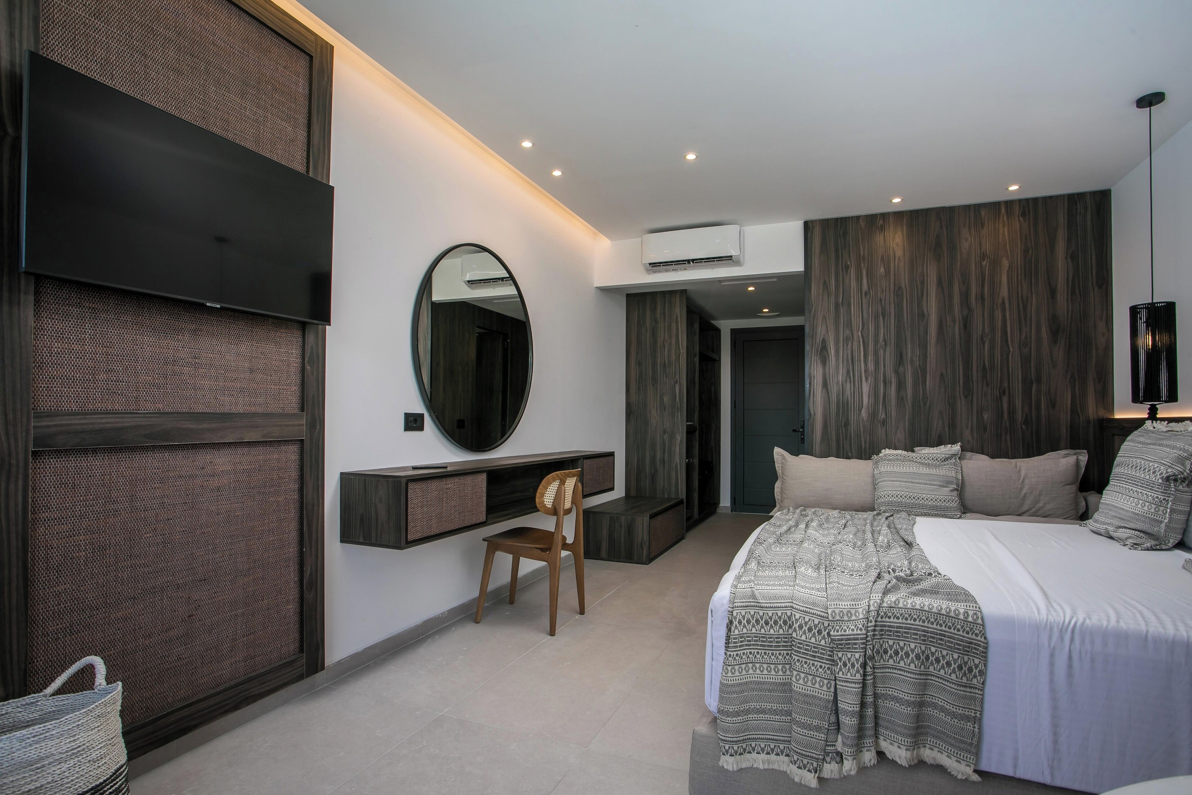 A modern bedroom with a large bed, a TV mounted on the wall, a desk with a chair, and an air conditioning unit.